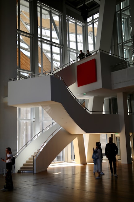 A modern architectural interior with large, angular windows allowing natural light to flood in. A central, curved staircase leads to an upper level, where two people stand looking out over the space. Below, three people are walking and interacting in the open area. The design features clean lines and a prominent red square artwork on the wall.