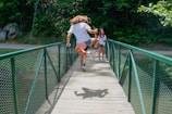 A person wearing a patriotic American flag-themed outfit is energetically jumping on a narrow green metal bridge surrounded by lush greenery. A friend in casual summer attire watches and smiles with a drink in hand.
