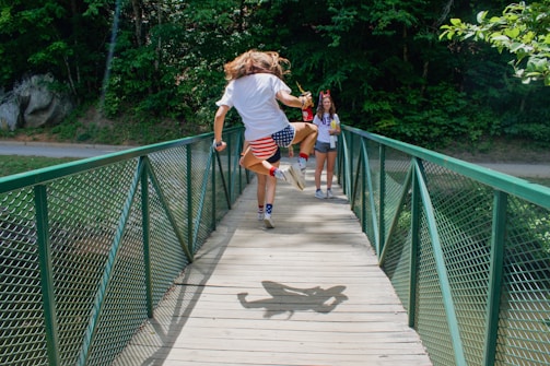 A person wearing a patriotic American flag-themed outfit is energetically jumping on a narrow green metal bridge surrounded by lush greenery. A friend in casual summer attire watches and smiles with a drink in hand.