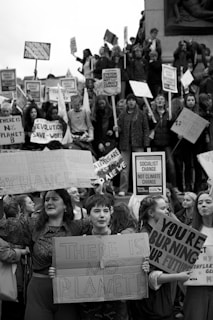 A black and white image of a large group of people, primarily young adults, holding protest signs. The crowd appears to be part of a climate change demonstration, as the signs carry messages such as 'There is no Planet B' and 'You're burning our future.' The group is gathered tightly together, showing solidarity for the cause.