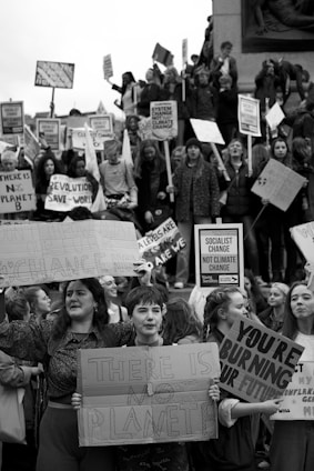 A black and white image of a large group of people, primarily young adults, holding protest signs. The crowd appears to be part of a climate change demonstration, as the signs carry messages such as 'There is no Planet B' and 'You're burning our future.' The group is gathered tightly together, showing solidarity for the cause.