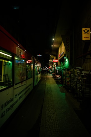 A nighttime urban scene with a tram moving along city streets. The area is dimly lit, with neon signs and streetlights providing illumination. There's a sidewalk alongside the tram with a newsstand displaying magazines or newspapers. A few people are visible in the background, suggesting a lively urban environment.