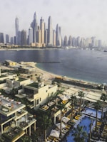 A cityscape with numerous modern skyscrapers overlooking a coastal area. In the foreground, there are stylish buildings with outdoor patios and pools, surrounded by palm trees and shaded areas with lounge chairs. A sandy beach extends along the shoreline, with a few people visible enjoying the water and sun.