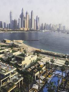 A cityscape with numerous modern skyscrapers overlooking a coastal area. In the foreground, there are stylish buildings with outdoor patios and pools, surrounded by palm trees and shaded areas with lounge chairs. A sandy beach extends along the shoreline, with a few people visible enjoying the water and sun.