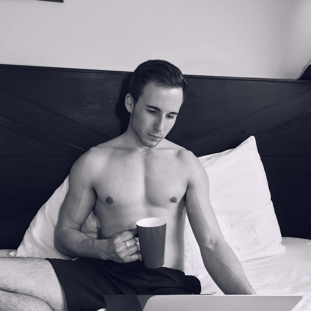 black and white photo of man holding mug while using laptop computer on bed