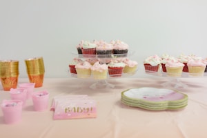 Cupcakes with pink and white frosting are arranged on tiered trays. Stacks of golden cups and smaller pink cups contain candies. Pink napkins with 'Oh Baby!' text are placed next to the cups, suggesting a baby shower theme. A set of pastel-colored plates rests nearby.