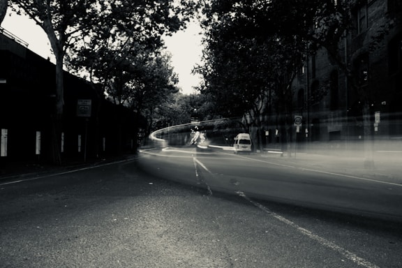 A long exposure photograph of an urban street at night, featuring blurred light trails from passing vehicles. The scene is framed by tall trees and brick buildings, with dim street lighting casting shadows.