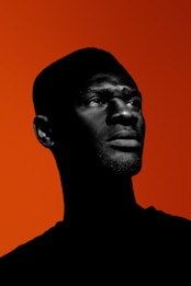 A silhouette of a person is depicted against a vibrant orange background. The contrast between the dark figure and the bright backdrop creates a striking visual. The person's facial features are highlighted by the light, giving a dramatic and bold impression.