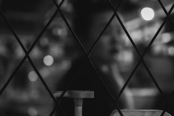 A black and white image featuring a blurred figure in the background, viewed through a diamond-patterned wire mesh. The foreground shows a pump dispenser slightly out of focus. The lighting creates soft bokeh effects, contributing to an abstract and mysterious atmosphere.