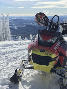 A snowmobile is resting on a snowy landscape with a scenic mountainous background. The snowmobile is primarily red and yellow with visible branding, and a helmet is placed on the handlebars. The snow-covered trees and expansive view create a winter adventure atmosphere.