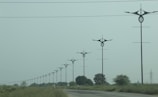 A series of uniquely designed power poles are aligned along a road surrounded by green grass and sparse trees under an overcast sky. The poles have distinctive, modern shapes that differentiate them from traditional utility poles.