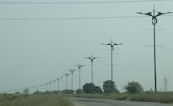 A series of uniquely designed power poles are aligned along a road surrounded by green grass and sparse trees under an overcast sky. The poles have distinctive, modern shapes that differentiate them from traditional utility poles.