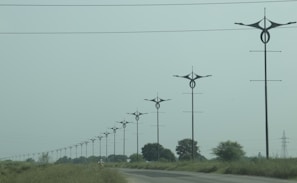 A series of uniquely designed power poles are aligned along a road surrounded by green grass and sparse trees under an overcast sky. The poles have distinctive, modern shapes that differentiate them from traditional utility poles.