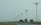 A series of uniquely designed power poles are aligned along a road surrounded by green grass and sparse trees under an overcast sky. The poles have distinctive, modern shapes that differentiate them from traditional utility poles.