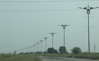 A series of uniquely designed power poles are aligned along a road surrounded by green grass and sparse trees under an overcast sky. The poles have distinctive, modern shapes that differentiate them from traditional utility poles.