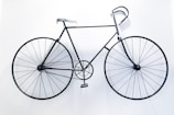 A black metal bicycle is mounted on a stark white wall, appearing almost like a minimalist sculpture. The design is simplistic with a thin frame and large circular wheels that emphasize a sense of balance and symmetry.