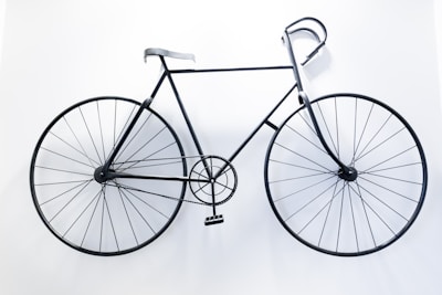 A black metal bicycle is mounted on a stark white wall, appearing almost like a minimalist sculpture. The design is simplistic with a thin frame and large circular wheels that emphasize a sense of balance and symmetry.