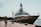 A luxurious yacht docked at a harbor alongside a stone paved walkway with pedestrians strolling nearby. The scene is set against a backdrop of clear skies and calm waters, with lampposts lining the path. The yacht is large and sleek, suggesting luxury and grandeur.