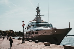 A luxurious yacht docked at a harbor alongside a stone paved walkway with pedestrians strolling nearby. The scene is set against a backdrop of clear skies and calm waters, with lampposts lining the path. The yacht is large and sleek, suggesting luxury and grandeur.