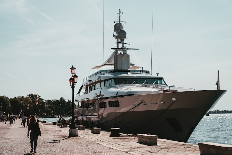 A luxurious yacht docked at a harbor alongside a stone paved walkway with pedestrians strolling nearby. The scene is set against a backdrop of clear skies and calm waters, with lampposts lining the path. The yacht is large and sleek, suggesting luxury and grandeur.