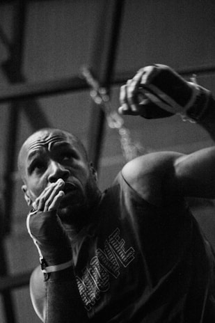 A person with wrapped hands appears to be in a training or fighting stance, raising one arm as if preparing to throw a punch. The scene is in black and white, and the setting appears to be indoors with a somewhat gritty or serious atmosphere.