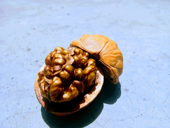 A close-up view of an opened walnut shell resting on a light blue surface. The walnut inside is golden brown and textured, casting a shadow.