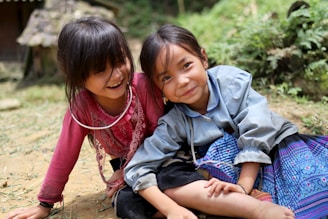 Two young children are sitting outdoors on the ground, dressed in colorful traditional clothing. One child is wearing a pink top and laughing, while the other child in a blue top is smiling warmly. The background features a blurred mix of greenery and a rustic structure.