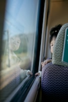 A young child is leaning on the back of a seat, gazing out the window of a moving vehicle. The reflection of their face is visible on the glass, and their hand lightly touches the window. The scene evokes a sense of curiosity and contemplation.