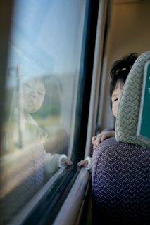 A young child is leaning on the back of a seat, gazing out the window of a moving vehicle. The reflection of their face is visible on the glass, and their hand lightly touches the window. The scene evokes a sense of curiosity and contemplation.