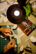 Hands are crafting paper models on a desk, surrounded by a variety of tools and materials. A lamp illuminates the workspace, and there are several sheets of paper, a glue stick, and a roll of tape. A green cutting mat and some green plants add color to the otherwise neutral space.