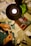 Hands are crafting paper models on a desk, surrounded by a variety of tools and materials. A lamp illuminates the workspace, and there are several sheets of paper, a glue stick, and a roll of tape. A green cutting mat and some green plants add color to the otherwise neutral space.
