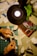 Hands are crafting paper models on a desk, surrounded by a variety of tools and materials. A lamp illuminates the workspace, and there are several sheets of paper, a glue stick, and a roll of tape. A green cutting mat and some green plants add color to the otherwise neutral space.