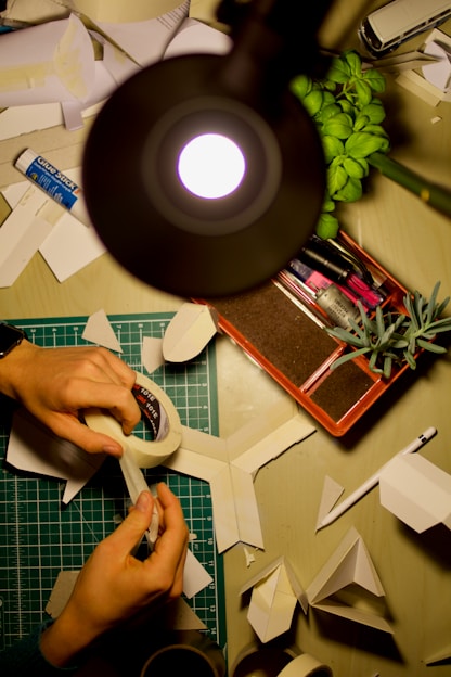 Hands are crafting paper models on a desk, surrounded by a variety of tools and materials. A lamp illuminates the workspace, and there are several sheets of paper, a glue stick, and a roll of tape. A green cutting mat and some green plants add color to the otherwise neutral space.