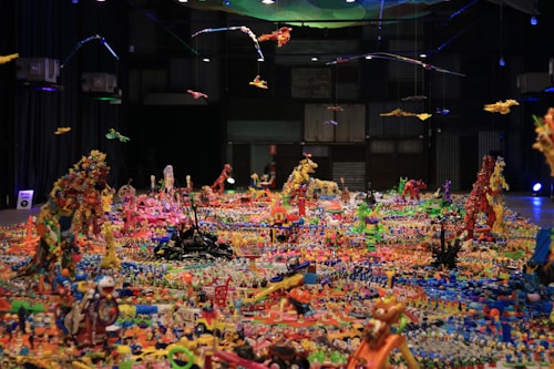 A large, intricate installation made up of numerous colorful toys and objects arranged on the floor. The scene is bright and vivid, featuring structures built from toys, with some suspended from above. The background is dark, with industrial elements like metal panels and lights adding contrast to the vibrant chaos on the floor.