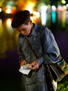 An elderly person wearing a beret and a denim jacket stands against a backdrop of colorful, out-of-focus city lights that reflect on water. They are focused intently on something they are holding, which appears to be a piece of paper or a ticket, in their hands. A strap of a bag with multicolored patterns is slung across the shoulder.