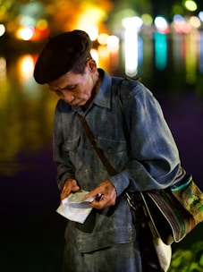 An elderly person wearing a beret and a denim jacket stands against a backdrop of colorful, out-of-focus city lights that reflect on water. They are focused intently on something they are holding, which appears to be a piece of paper or a ticket, in their hands. A strap of a bag with multicolored patterns is slung across the shoulder.