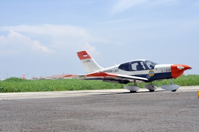 A small aircraft parked on a runway with a clear blue sky above and green grass in the background. The aircraft is white with some red and orange accents, displaying the number L-210. The foreground features a paved surface, and the aircraft seems to belong to an aviation organization.