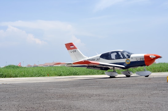 A small aircraft parked on a runway with a clear blue sky above and green grass in the background. The aircraft is white with some red and orange accents, displaying the number L-210. The foreground features a paved surface, and the aircraft seems to belong to an aviation organization.