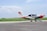 A small aircraft parked on a runway with a clear blue sky above and green grass in the background. The aircraft is white with some red and orange accents, displaying the number L-210. The foreground features a paved surface, and the aircraft seems to belong to an aviation organization.