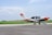 A small aircraft parked on a runway with a clear blue sky above and green grass in the background. The aircraft is white with some red and orange accents, displaying the number L-210. The foreground features a paved surface, and the aircraft seems to belong to an aviation organization.