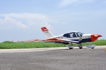 A small aircraft parked on a runway with a clear blue sky above and green grass in the background. The aircraft is white with some red and orange accents, displaying the number L-210. The foreground features a paved surface, and the aircraft seems to belong to an aviation organization.