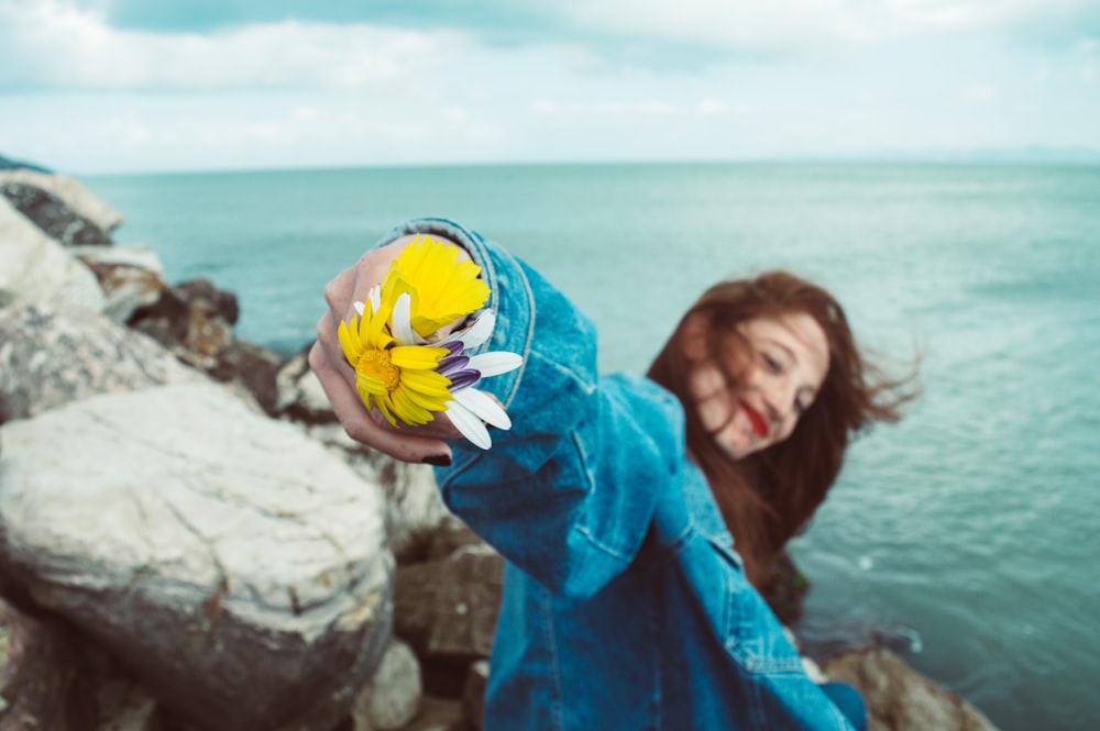 smiling woman wearing blue denim jacket holding yellow and white flower