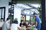A group of people gathered around an outdoor table, engaged in conversation and laughter. The setting appears to be a casual dining or cafe environment with a modern design. There are plants around the seating area and the atmosphere seems lively and friendly.