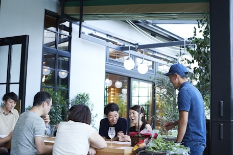 A group of people gathered around an outdoor table, engaged in conversation and laughter. The setting appears to be a casual dining or cafe environment with a modern design. There are plants around the seating area and the atmosphere seems lively and friendly.