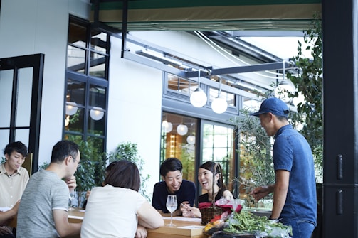 A group of people gathered around an outdoor table, engaged in conversation and laughter. The setting appears to be a casual dining or cafe environment with a modern design. There are plants around the seating area and the atmosphere seems lively and friendly.
