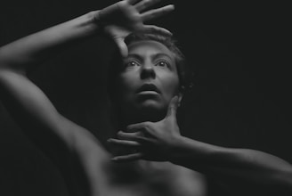 A black and white portrait of a person with their hands positioned around their face, creating a dramatic and expressive look. The lighting emphasizes the contours of the face and body, casting deep shadows that add depth and intrigue.