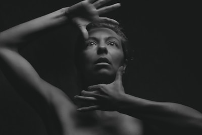 A black and white portrait of a person with their hands positioned around their face, creating a dramatic and expressive look. The lighting emphasizes the contours of the face and body, casting deep shadows that add depth and intrigue.