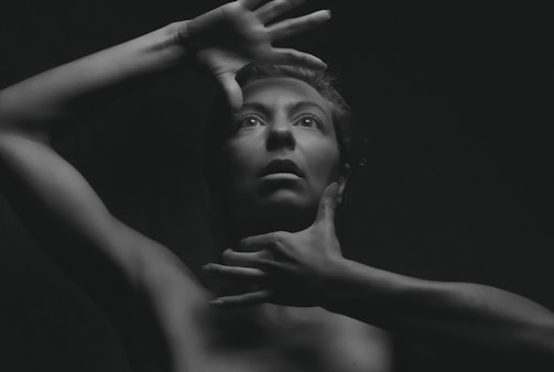 A black and white portrait of a person with their hands positioned around their face, creating a dramatic and expressive look. The lighting emphasizes the contours of the face and body, casting deep shadows that add depth and intrigue.