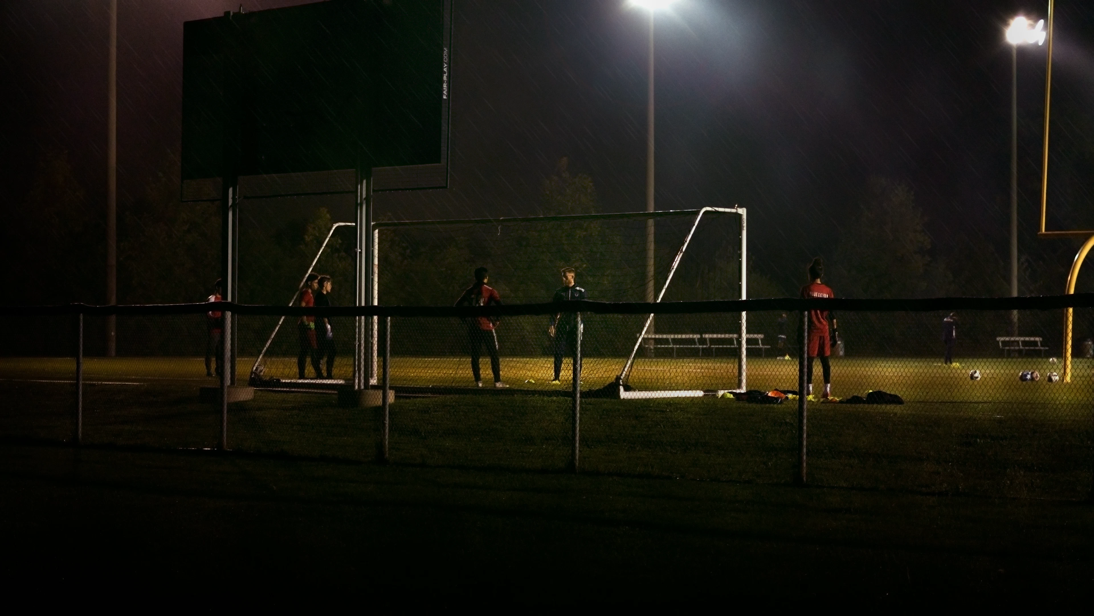 Goal frames under a floodlight at night, players waiting at the side of the pitch