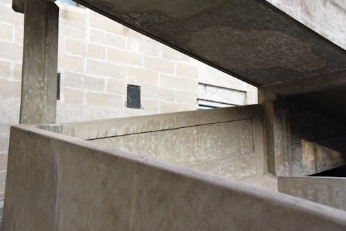 A concrete structure featuring angular lines and geometric shapes, with a textured brick wall in the background. The play of light and shadows emphasizes the architecture's industrial aesthetic.
